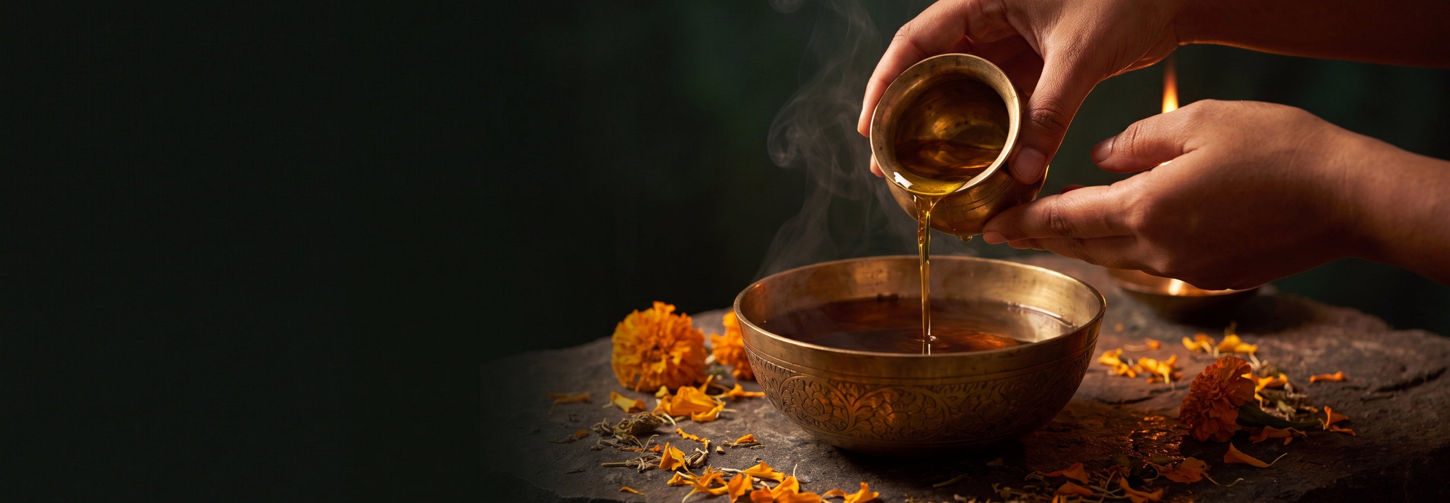 Hands pouring golden Ayurvedic oil into a brass bowl by lamplight, with marigold petals on dark stone